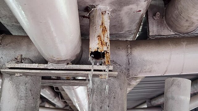 Rusty industrial pipe leaks water from corroded joint beneath metal structure, showing infrastructure damage, plumbing failure and maintenance issues.