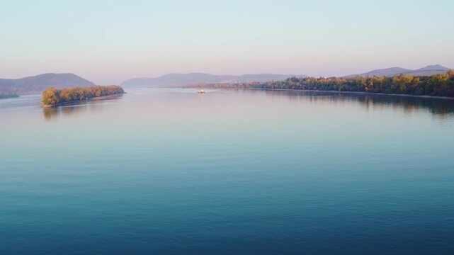 Wide scenic shot of the peaceful Danube river flowing gently, featuring a small wooded island, distant hills, and a small boat navigating the calm blue waters at sunrise.