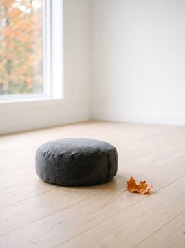 Minimalist meditation cushion on light wood floor, charcoal grey zafu in bright zen room, round floor pillow for yoga and mindfulness, autumn leaf in serene interior space.