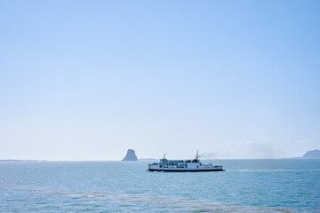 Passenger ferry glides across calm blue ocean toward a rocky island on a clear sunny day, evoking coastal travel, maritime transport and peaceful open-water scenery ideal for travel and nature themes. © Loginova