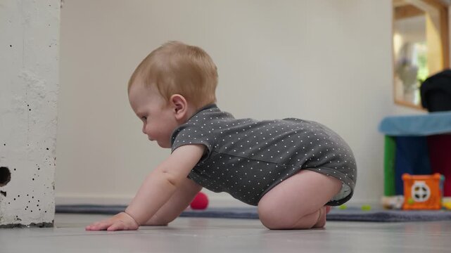Infant in a dark gray dotted onesie gets on all fours and sits on the floor in the nursery near a white textured column, lit by soft indoor light from the right