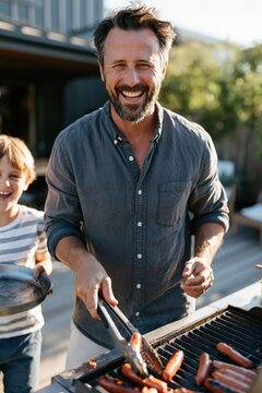Bearded man grilling sausages at backyard barbecue with smiling child for summer banner and family gathering background