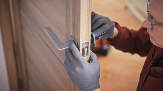A specialist in gloves and uniform fixes a door lock mechanism on a wooden interior door with an orange toolbox visible in the background. Concept of home repair and door maintenance