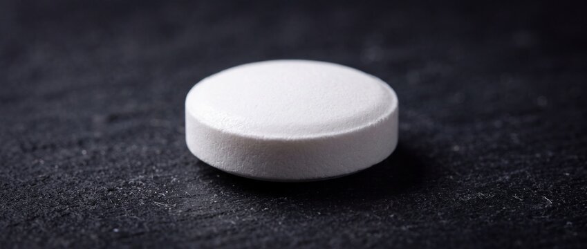 Single round white tablet pill placed centrally on dark textured background representing pharmacy medication medical treatment health care wellness nutrition theme.