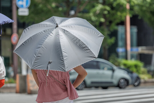 Candid shot of a woman in a red gingham blouse and white pants holding a silver, scalloped frilled parasol at a busy city crossing.