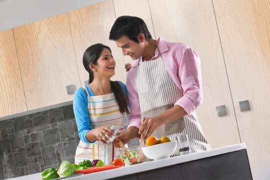 Happy Indian couple adding salt to fresh salad in kitchen