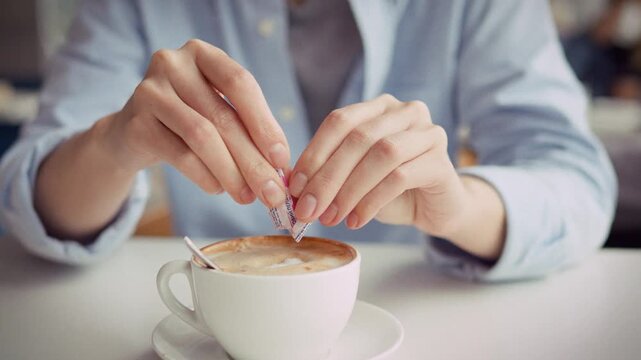 Girl's hands sprinkle sugar from packet into a fresh cappuccino at a daytime roadside cafe in the bright morning light, the coffee steaming on a small table as she sits, adding sweetness to the drink