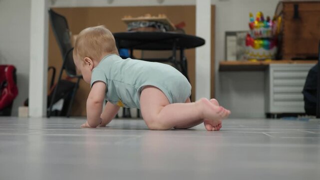 A baby in a light blue onesie is trying to crawl on all fours to the left on a gray floor in a room at chest and eye level. Infant tries to crawl