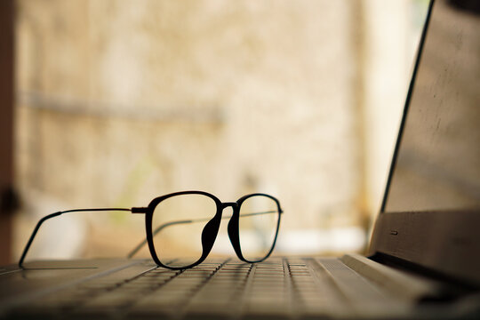 Close up of stylish black frame eyeglasses resting on a laptop keyboard in a warm bright workspace
