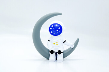 Miniature astronaut toy with blue starry helmet sitting on gray crescent moon on white background. Childrens space education, STEM learning toy for science popularization, dream, imagination concept. © Ninaveter