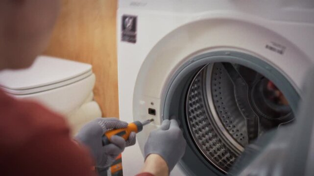 A close-up of gloved hands unscrewing bolts from the body of a front-loading washing machine in a bathroom. Concept of disassembling and repairing a front-loading washing machine at home