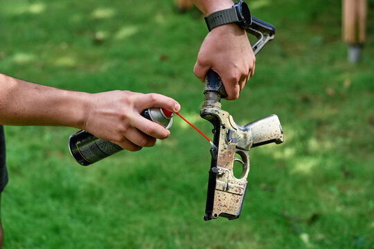 Close-up of hands spraying lubricant into a AR rifle lower while performing outdoor maintenance on grass. The image conveys repair work, care,