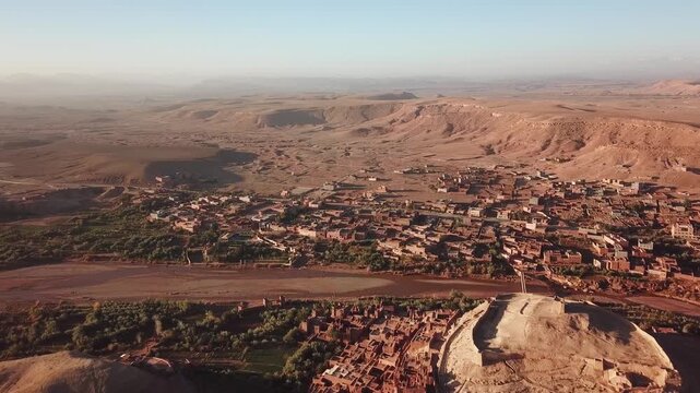 Aerial view of the historic earthen clay architecture of the Kasbah of Ait Benhaddou, surrounded by arid desert landscape and a dry riverbed in Ait Benhaddou, Draa-Tafilalet, Morocco.
