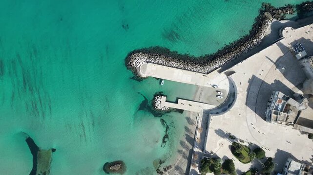 Aerial view of the turquoise waters, stone pier, and small boats along the coastal promenade in Otranto, Apulia, Italy.