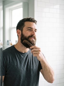 Handsome man brushing his thick beard with a wooden comb in a bright bathroom showing male grooming routines personal hygiene and No Shave November awareness