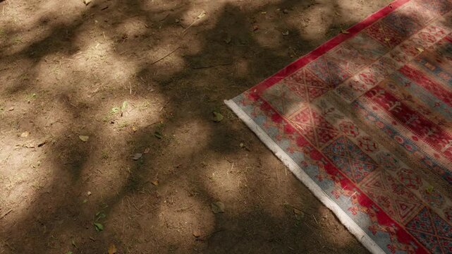 Edge of a red patterned rug laid on dry bare earth under dappled tree shade