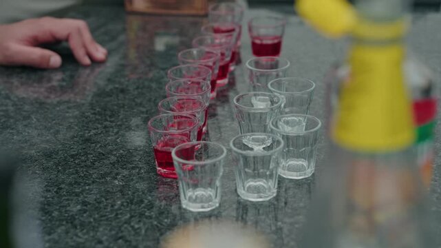 Two rows of small glasses with red drink and empty glasses lined up on a granite bar