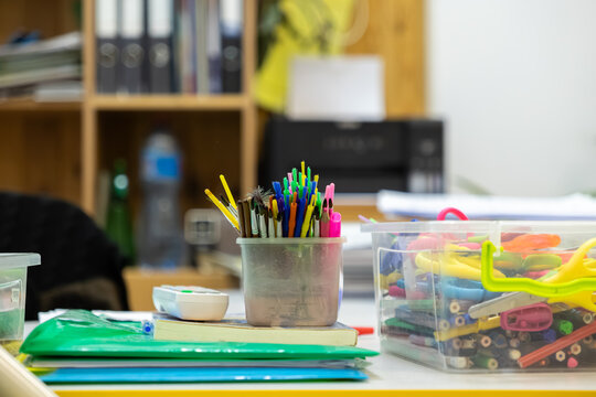 Colorful paintbrushes in a plastic cup and office supplies inside a container on a messy desk, representing a creative workspace for art projects in a school or home classroom