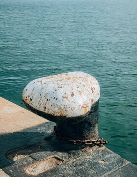 Close up view of a rusty weathered mooring bollard with iron chain at a harbor dock against calm blue sea water.