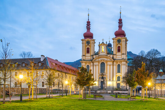 The former Franciscan monastery and Church of the Visitation stand tall in Hejnice during the evening. Lights illuminate the area as trees and buildings surround the site.