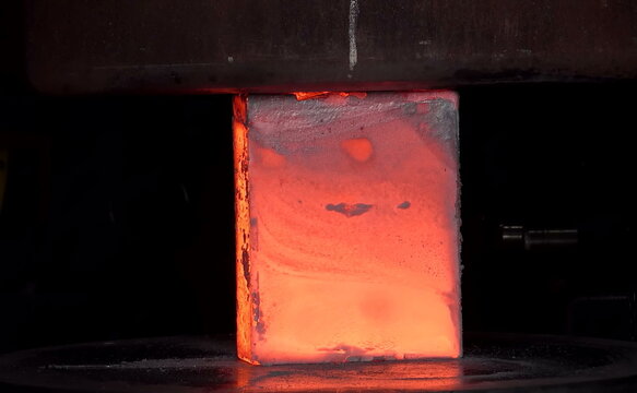 A heated cylindrical metal block glows bright orange as sparks and fragments scatter under compression, showing the intensity and precision of industrial forging at extreme temperature levels.  