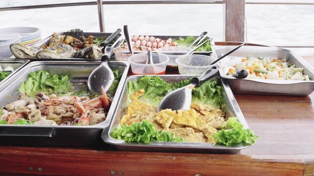 Buffet seafood spread on a wooden table aboard a boat with sea view. Includes grilled fish, prawns, salad, and mixed vegetables in stainless trays. Tropical cruise dining and catering concept.