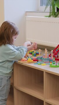 Subject stands by radiator playing stackable items. Visible red string held in her right hand during play. Wooden table has toys including rings on wooden pegs and blocks