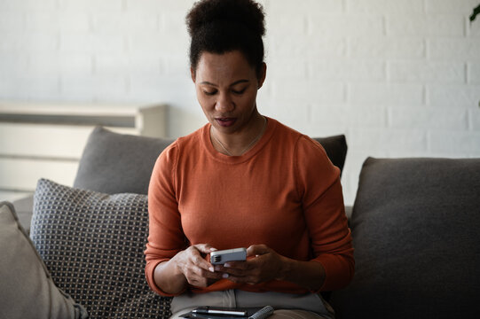 Focused woman uses a smartphone while sitting on a sofa at home, checking messages, information or online services during her daily routine