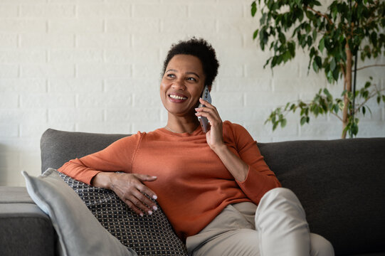 Happy woman smiles while talking on a smartphone and relaxing on a sofa at home, enjoying a pleasant phone conversation or good news