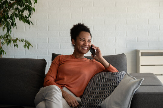 Cheerful woman has a friendly smartphone conversation while lounging on a sofa at home, smiling and enjoying the call