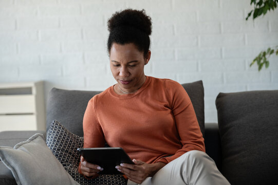 Mature woman using a digital tablet while sitting comfortably on a sofa at home, focused on online information