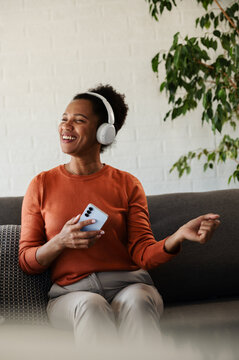 Happy woman wearing wireless headphones smiles and moves to music while holding a smartphone and relaxing on a sofa at home