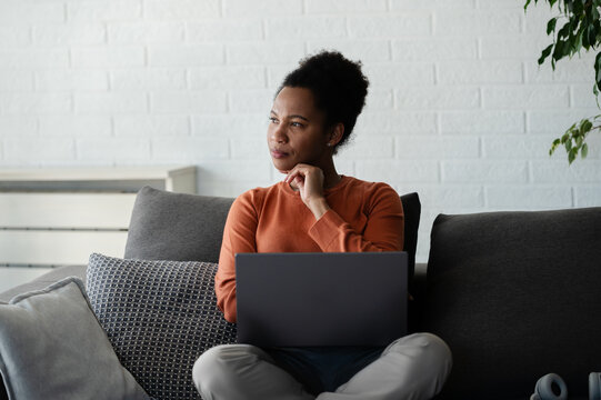 Thoughtful woman sits on a sofa with a laptop, looking away while thinking, planning or considering a problem at home.
