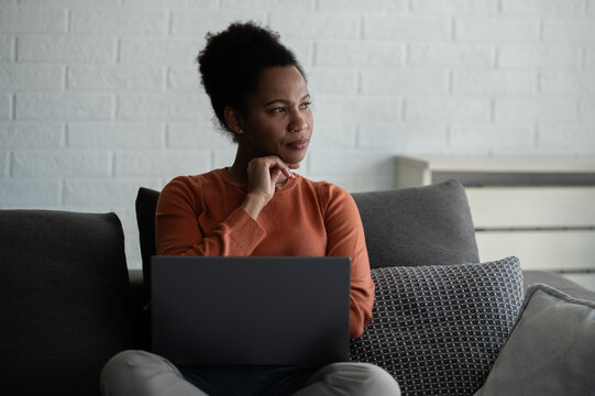 Professional woman pauses to think while working on a laptop from her sofa at home, representing remote work, planning and decision making