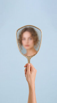Hand holding mirror with blurred female reflection, light blue background. Minimal concepts of self perception, identity, anonymity, uncertainty, difference between appearance and personal perception.