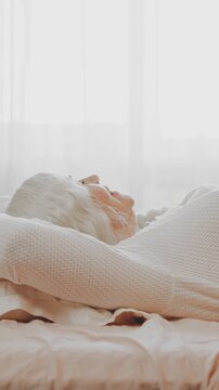 Happy senior elderly woman falls on soft pillow to rest lunchtime afternoon nap, good sleep in cozy room, orthopedic mattress cotton fabric bed linen. bedroom day sun light from window. lady old age.