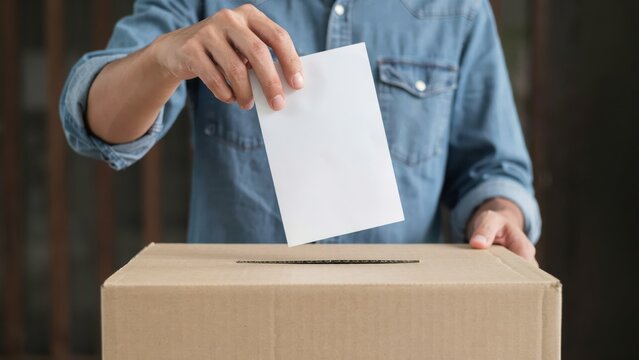 Person casting a vote by dropping a white ballot into a cardboard election box during an election.