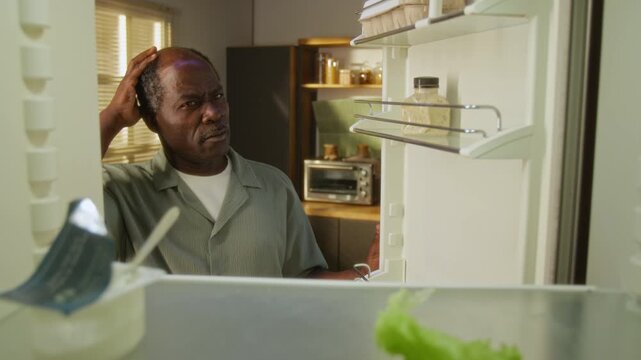 Senior African American man opening empty refrigerator, scratching head in confusion and inspecting single jar. Footage is perfect for grocery delivery services or food insecurity campaigns