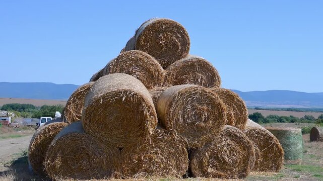 A slow zoom-out reveals a large pyramid stack of round hay bales in a vast agricultural field, with a tractor, farm building, and mountains in the background.