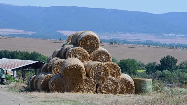 a large pyramid stack of round hay bales in a vast agricultural field, with a tractor, farm building, and mountains in the background.