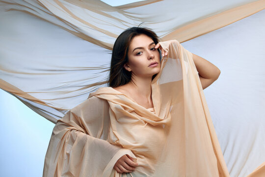Elegant woman wrapped in flowing cream fabric against airy studio background. Concept of skincare branding, wellness ads and resort fashion.