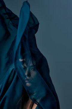Artistic beauty portrait of woman seen through translucent dark teal fabric. Concept of perfume branding, conceptual beauty and fashion editorial.