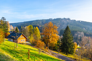 Obraz premium Autumn showcases colorful trees in Albrechtice, Jizera Mountains, Czechia. A wooden house sits on a hillside with mountains in the background and a clear sky above.