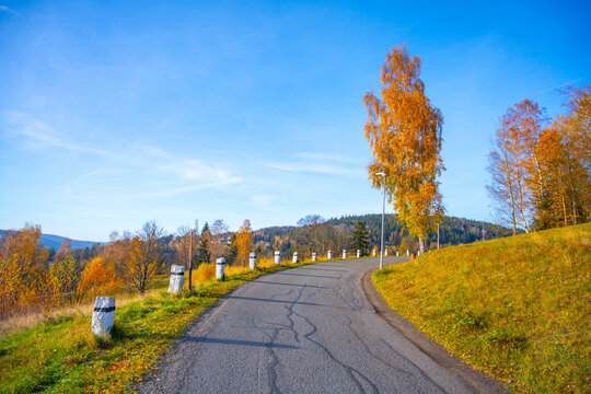 The winding road in Albrechtice shows bright autumn colors from trees in the Jizera Mountains. The landscape features a clear sky and vibrant foliage.