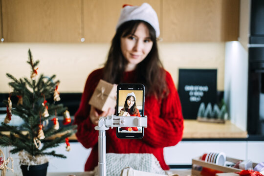 Woman in Santa hat shows wrapped kraft box to phone on tripod in decorated kitchen. Christmas content creator kitchen, christmas DIY content filming, christmas craft video creation