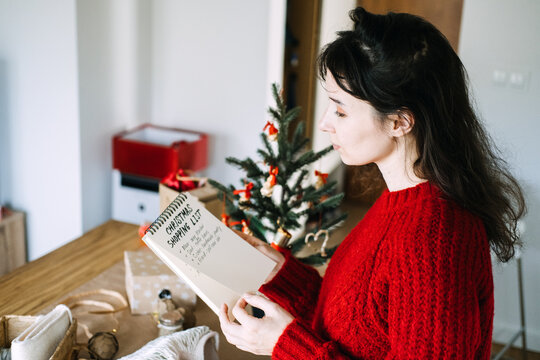 Woman turns page of notebook with CHRISTMAS SHOPPING LIST beside wrapped gifts and small tree. Personalized christmas gift list, gift ideas for each family member, thoughtful christmas gift planning