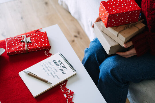 Woman holds stack of wrapped gifts beside notebook with HOLIDAY BUDGET PLAN on table. Christmas budget planning, mindful holiday spending, no impulse buying gift rules