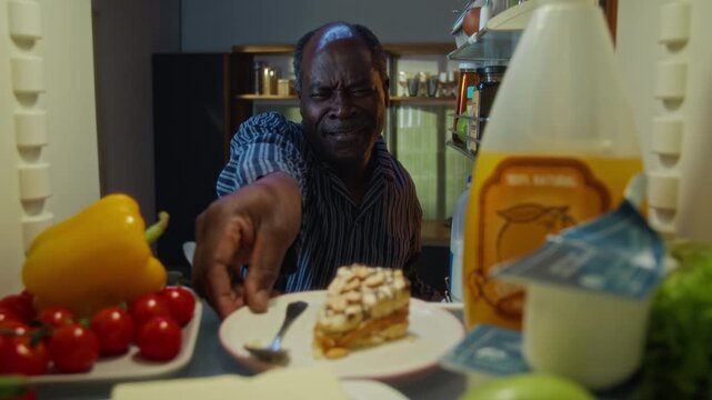 Senior African American man opening refrigerator, looking inside hesitantly, and taking out plate with slice of cake. Footage represents concepts of late-night snacking or diet temptation