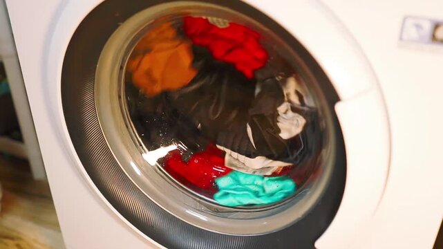 Close up of a modern white washing machine with a rotating glass drum filled with bright laundry spinning at home or laundromat.