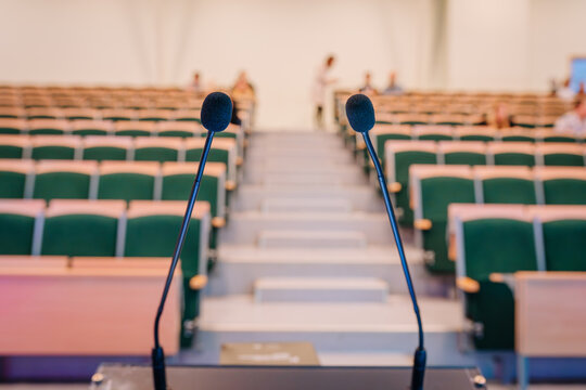 Two microphones positioned on a podium facing empty rows of green seating in a modern auditorium with bright lighting and a spacious layout